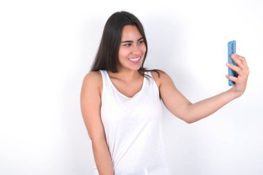 Beyazın üzerine beyaz bluz giyen neşeli, güzel esmer kadının izole edilmiş fotoğrafı, cep telefonuyla selfie çekiyor. İnsanlar, teknoloji ve eğlence kavramı