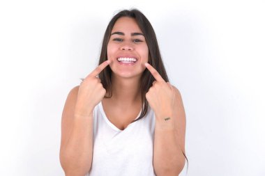 Güçlü, sağlıklı, beyaz dişler. Beyaz üzerine beyaz bluz giyen mutlu esmer kadının mükemmel beyaz dişleri işaret eden parlak gülümsemesiyle yakın plan portresi..