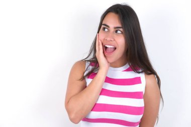 Güzel esmer kadın beyaz duvarın üzerinde çizgili bluz giyiyor. Heyecanlı bir şekilde yüzüne bakıyor. Reklam ve şaşkınlık konsepti.