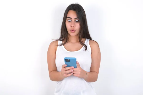 白い目に白い上に白いトップを身に着けている美しいブルネットの女性は 現代のスマートフォンを保持し 友人からの予期しないメッセージを受け取ります リマインダーを読む — ストック写真