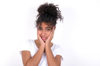 Üzgün kız iki eliyle yüzüne dokunuyor. Beyaz tişört giyen, beyaz duvarın üzerinde poz veren güzel, melez bir kadın.