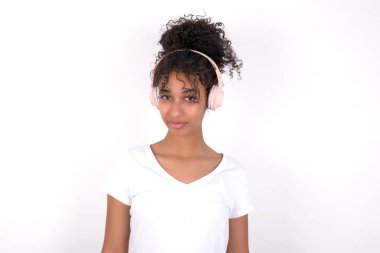 Kulaklık takan kafası karışmış bir kız. Beyaz tişört giyen, beyaz duvarın üzerinde poz veren güzel, melez bir kadın.