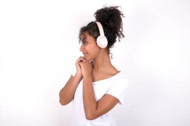 Kız müzik kulaklığı takıyor. Konsantre müzik dinliyor ve ilgiyle yan tarafa bakıyor. Beyaz tişört giyen, beyaz duvarın üzerinde poz veren güzel, melez bir kadın.