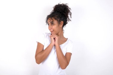 Meraklı kız ellerini çenesinin altında tutuyor dudaklarını ısırıyor ve ilgiyle bakıyor. Beyaz tişört giyen, beyaz duvarın üzerinde poz veren güzel, melez bir kadın.