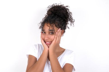 Mutlu kız her iki yanağına nazikçe dokunur, şefkatli bir gülümsemesi vardır, beyaz dişlerini gösterir, kameraya doğru dik dik bakar. Beyaz tişört giyen, beyaz duvarın üzerinde poz veren güzel, melez bir kadın.