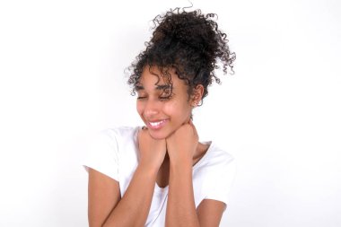 Neşeli kızın utangaç, mutlu bir yüzü var. Gülümsüyor, beyaz dişleri var. Beyaz tişört giyen, beyaz duvarın üzerinde poz veren güzel, melez bir kadın.