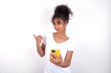 Kız başparmağıyla işaret ediyor ve boş alan gösteriyor. Cep telefonunu mesaj göndermek için tutuyor. Beyaz tişört giyen, beyaz duvarın üzerinde poz veren güzel, melez bir kadın.