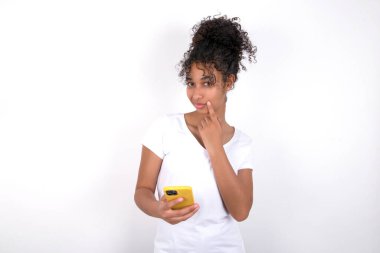 Kız bir şey hakkında derin düşünüyor, modern cep telefonu kullanıyor, iyi bir mesaj uydurmaya çalışıyor, işaret parmağını dudaklarının yakınında tutuyor. Beyaz tişört giyen, beyaz duvarın üzerinde poz veren güzel, melez bir kadın.