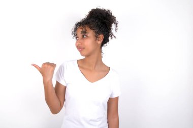 Kız işaret eder ve tavsiye verir. Reklam yapar. Beyaz tişört giyen, beyaz duvarın üzerinde poz veren güzel, melez bir kadın.