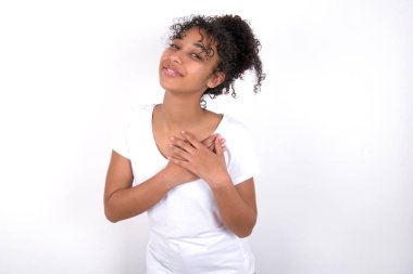 Beyaz arka planda beyaz tişört giyen afro saçlı güzel bir kız. Mutluluğu ifade ediyor, güzel gülüyor, ellerini kalbinin üstünde tutuyor.