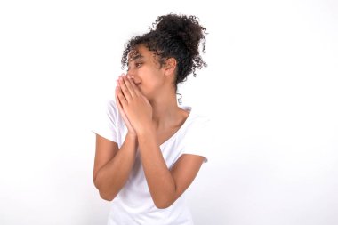 Coşkulu, genç, afro saçlı beyaz tişörtlü güzel bir kız beyaz arka planda neşeyle gülüyor ve avuç içlerini birbirine bastırıyor.