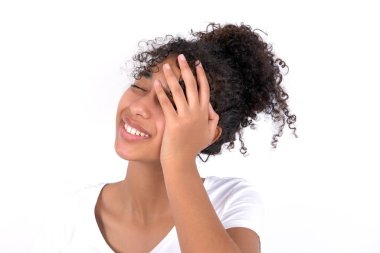 Beyaz arka planda beyaz tişörtlü, afro saçlı genç bir kız yüzü avuçlarına doluyor ve gülümsüyor, kıkırdıyor, komik pozlar duyuyor.