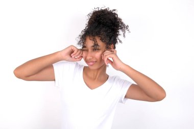 Beyaz arka planda beyaz tişört giyen mutlu, genç, güzel bir kız yüksek sesli müziği görmezden geliyor ve parmaklarıyla kulaklarını tıkıyor sesi kısmak istiyor.