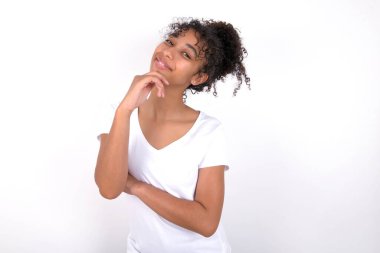 Beyaz arka planda beyaz tişörtlü, afro saçlı güzel bir kız mutlu bir şekilde gülüyor. Çenesinde elini tutuyor. Olumlu duygular ifade ediyor.