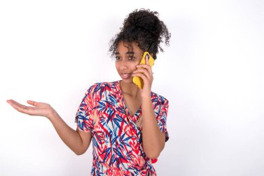 Telefonda konuşan kız, eli yüzü asık, utancından ve şaşırmış, sinirli ve hayal kırıklığına uğramış. Korku ve hata için üzüntü. Beyaz duvarın üzerinde duran renkli elbiseli güzel melez kadın.