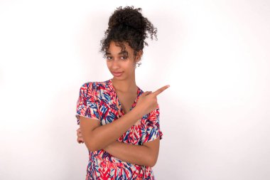 Kız kameraya geniş bir şekilde gülümsüyor, parmaklarını uzatıyor, ilginç ve heyecan verici bir şey gösteriyor. Beyaz duvarın üzerinde duran renkli elbiseli güzel melez kadın.