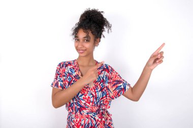 Işıl ışıl gülümseyen, iki parmağıyla işaret eden ve boş fotokopi alanına bakan pozitif kız. Reklam konsepti. Beyaz duvarda renkli elbiseler giyen güzel melez bir kadın.