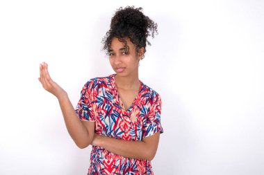 Studio shot of frustrated girl gesturing with raised palm, frowning, being displeased and confused with dumb question. Young beautiful mixed race woman wearing colourful dress over white wall