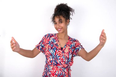 Neşeli kız kollarını başının üstüne kaldırarak hoş geldin jesti yapıyor. Beyaz duvarda renkli elbiseler giyen güzel melez bir kadın.