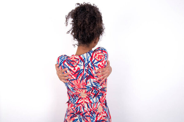 Girl hugging oneself happy and positive from backwards. Self love and self care. Young beautiful mixed race woman wearing colourful dress standing over white wall