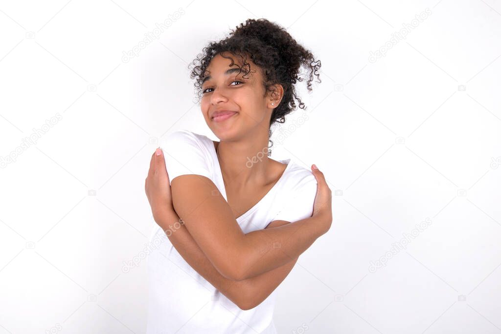 Charming pleased girl embraces own body, pleasantly feels comfortable poses. Tenderness and self esteem concept. Young beautiful mixed race woman wearing white t-shirt posing over white wall