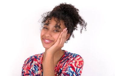 Gülümseyen mutlu kız tek eliyle yüzünü tutuyor. Beyaz duvarda renkli elbiseler giyen güzel melez bir kadın.