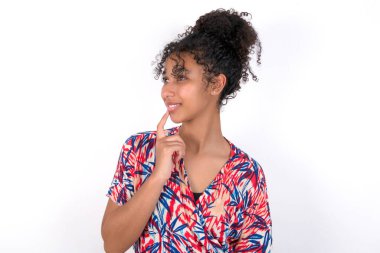 Kız rüya gibi görünüyor ve parmağını çenesine koyuyor. Beyaz duvarda renkli elbiseler giyen güzel melez bir kadın.