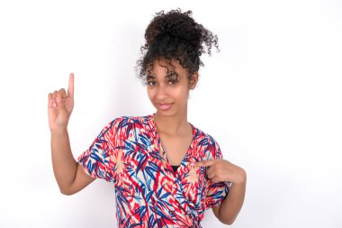 Kız bir elini diğer elini havaya kaldırarak işaret ediyor. Beyaz duvarda renkli elbiseler giyen güzel melez bir kadın.