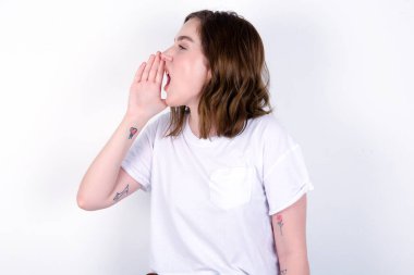 Kız profili, mutlu ve heyecanlı görünüyor, bağırıyor ve kopyalamak için arıyor. Beyaz tişörtlü beyaz bir kadın beyaz duvarın üzerinde poz veriyor.