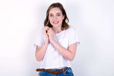 Hoş bir suratı olan rüya gibi çekici bir kız ellerini yüzüne çarpıyor, hoş bir şey için heyecanlanıyor. Beyaz tişörtlü beyaz bir kadın beyaz duvarın üzerinde poz veriyor.