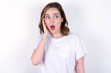 Beyaz arka planda beyaz tişört giyen beyaz tenli genç bir kadın heyecanla yüzüne bakıyor. Reklam ve şaşkınlık konsepti.
