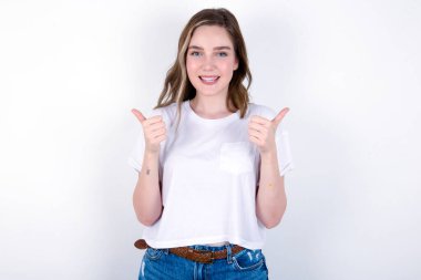 İyimser kız olumlu duygularla baş parmağını kaldırıyor. Kalite ve tavsiye konsepti. Beyaz arka planda beyaz tişört giyen beyaz bir kadın. 