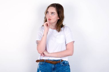 Elleri çenesinin altında ve yana bakan şüpheli ve şüpheci bir ifadeyle şüpheli ve şüpheci bir kız. Beyaz arka planda beyaz tişört giyen beyaz bir kadın. 