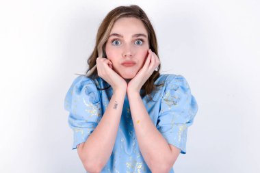 Şaşırmış suratlı bir kız ellerini çenesinin altında tutuyor dudaklarını katlıyor ve suratını ekşitiyor. Beyaz arka planda mavi tişört giyen beyaz bir kadın.