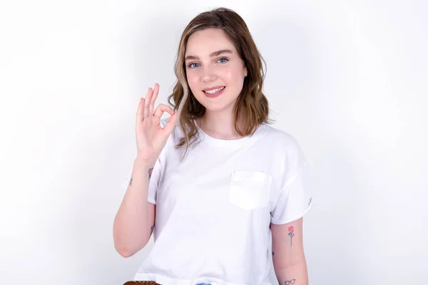 うれしい魅力的な少女は 承認を表すように手でOkのサインを示し 楽観的である陽気な表情を持っています 白い壁の上にポーズ白いTシャツを着ている若い美しい白人女性 — ストック写真