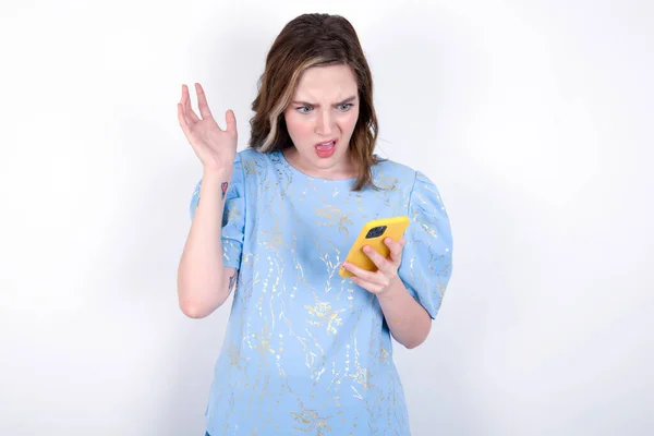Photo of outraged annoyed girl holds cell phone, makes call, argues with colleague,  expresses negative emotions. People and anger. Young caucasian woman wearing blue T-shirt over white background