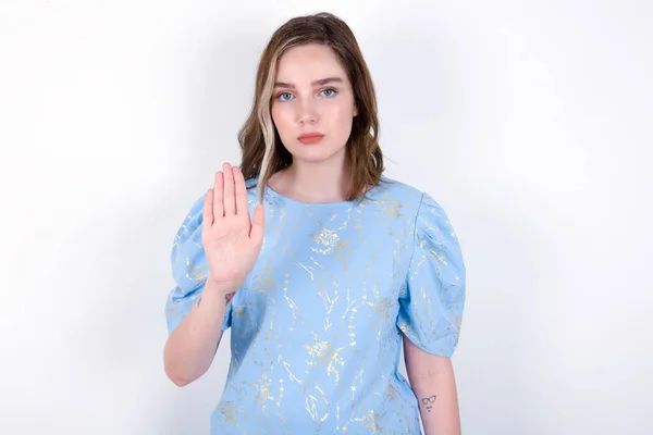 白い背景の上に青いTシャツを着ている若い白人女性は停止印 禁止シンボルを示します 女性は厳密な表現でカメラにパームを前進させます — ストック写真