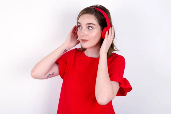 白い背景に赤いTシャツを着ている若い白人女性はステレオヘッドフォンを着て 音楽を脇に置いてリストします 人々の趣味のライフスタイルコンセプト — ストック写真
