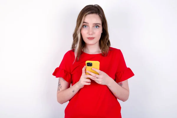 真面目な自信を持った若い白人女性の肖像画は 2つの手で携帯電話を保持する白い背景の上に赤いTシャツを着ています — ストック写真