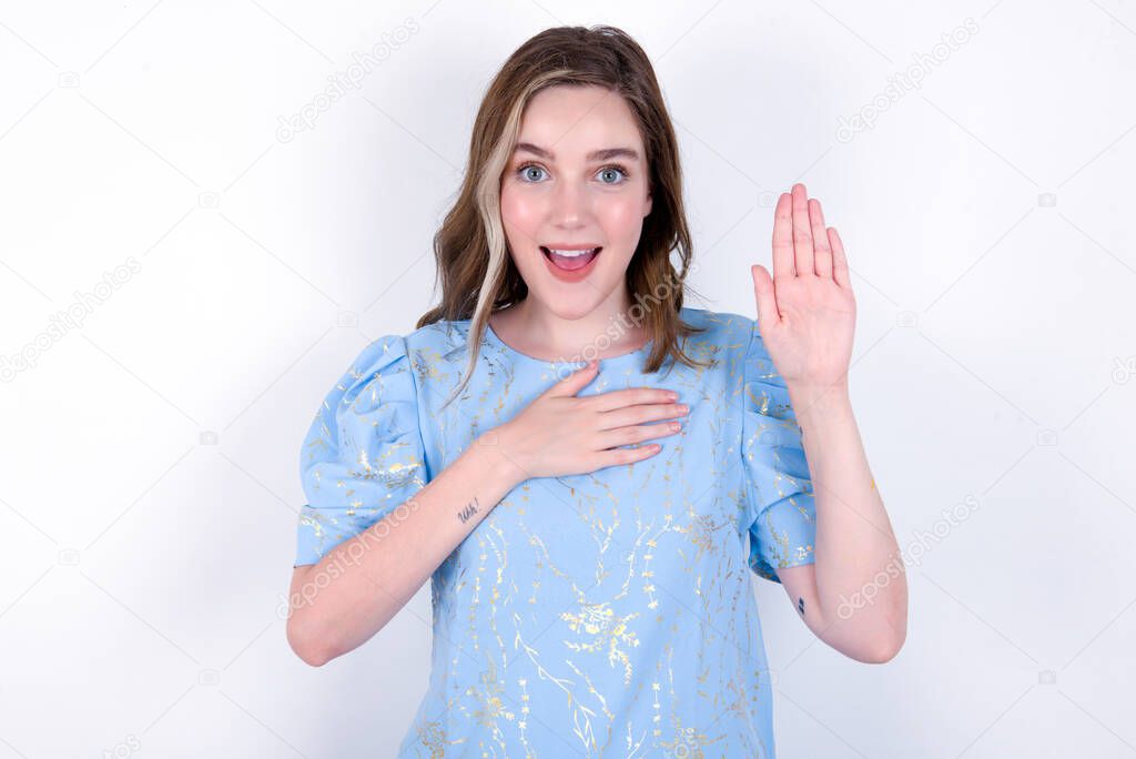 I swear, promise you not regret. Portrait of sincere young caucasian woman wearing blue T-shirt over white background raising one arm and hold hand on heart as give oath, telling truth, want you to believe.