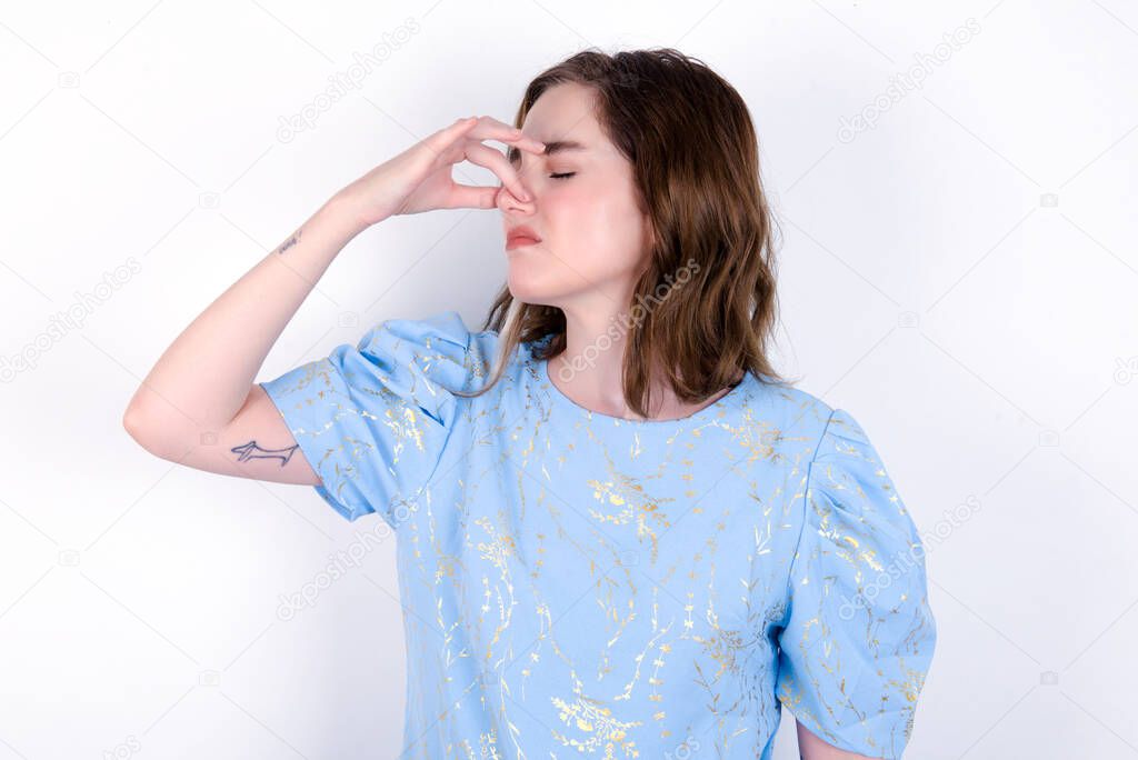 Displeased young caucasian woman wearing blue T-shirt over white background plugs nose as smells something stink and unpleasant, feels aversion, hates disgusting scent.