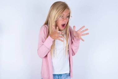 Beyaz arka planda pembe ceketli ve gözlüklü küçük sarışın kız şok edici hareketler yapıyor.