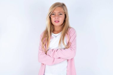 Pembe ceketli, beyaz arka planda gözlüklü, çapraz kollu küçük sarışın kız kameraya bakıyor.
