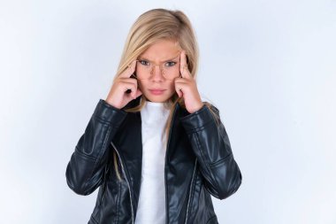 Beyaz arka planda motosiklet ceketi ve gözlük takan ciddi şekilde konsantre olmuş sarışın küçük kız parmaklarını şakaklarda tutuyor, gerginliği azaltmaya çalışıyor, düşüncelerle bir araya geliyor ve sınav için önemli bilgileri hatırlamaya çalışıyor.