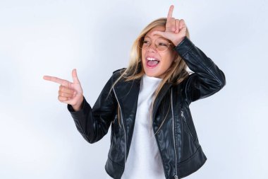 Beyaz arka planda motosiklet ceketi ve gözlük takan güzel sarışın kız ezik işareti yapıyor ve boş yeri işaret ediyor.