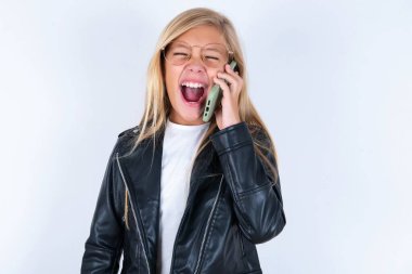 Aşırı duygusal, mutlu, sarışın, beyaz arka planda motosiklet ceketi ve gözlük takan küçük kız telefon görüşmeleri sırasında arkadaşından komik bir hikaye duyuyor.