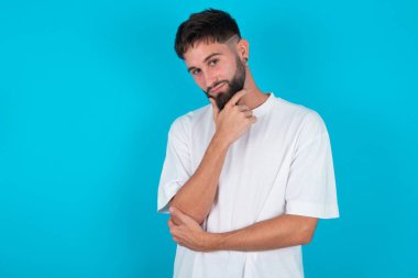 Mavi arka planda beyaz tişört giyen, sakallı, düşünceli beyaz adam elini çenesinin altında tutuyor, doğrudan kameraya bakıyor ve ilgi çekici bir şey dinliyor. Gençlik kavramı.