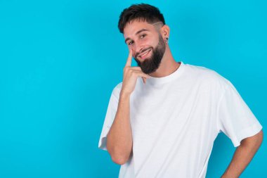 Beyaz tişörtlü beyaz sakallı beyaz bir adam mavi arka planda görünüyor, gözlemliyor, bir objeye öncülük ediyor ya da bir şeye dikkat ediyor..