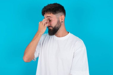 Çok üzgün sakallı, beyaz tişörtlü beyaz bir adam mavi arka planda burnuna dokunuyor, ağlamak istiyor, stresli bir ilişkisi var ya da işle ilgili sorunları var.