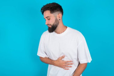 Genç, sakallı, beyaz tişörtlü, mavi arka planda, güçlü karın ağrısı çeken beyaz bir adam..